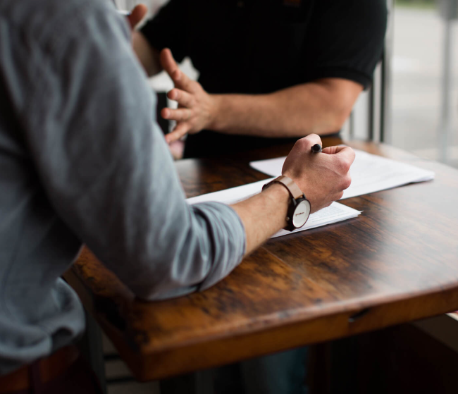 Two people are signing an important document while sitting at a wooden table.