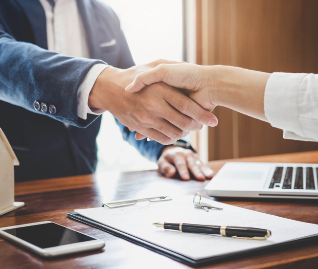 A picture of two people shaking hands over a desk with a laptop, keys, and pen on it.