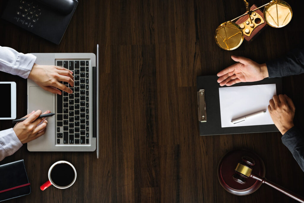 Two individuals at a desk with one using a laptop and the other reviewing documents, with legal paraphernalia