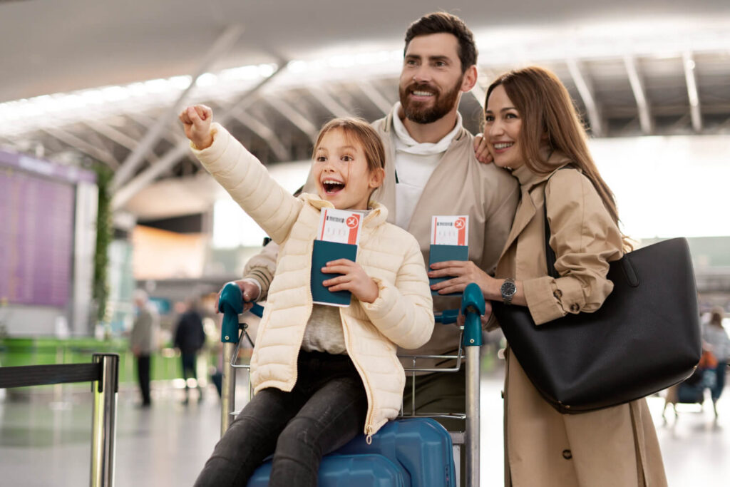 The picture shows a family of three at the airport. The little girl is sitting on top of the luggage cart, pointing at something excitedly. Her parents are standing next to her, smiling and looking at her. They all seem happy and excited.